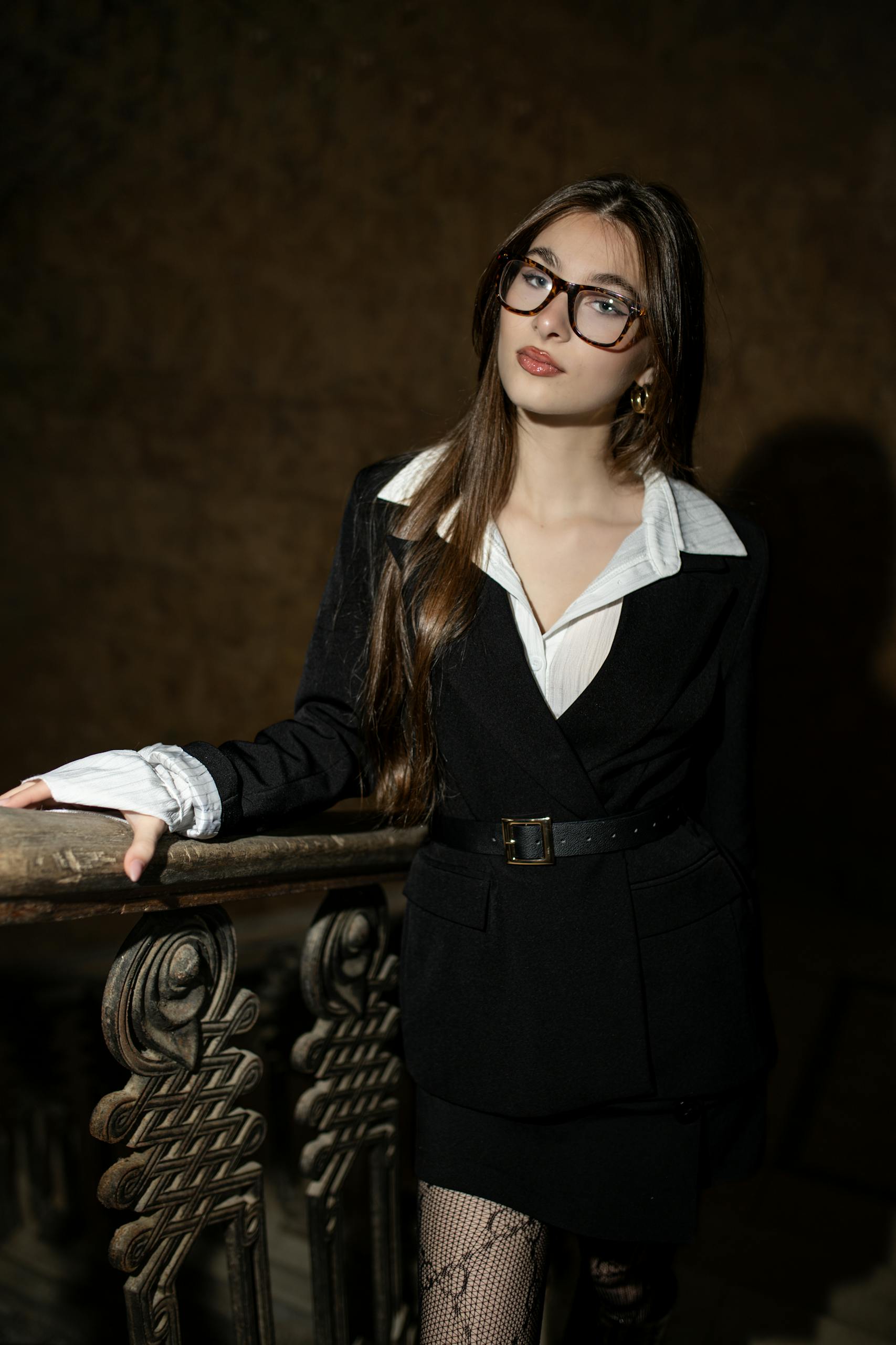 Stylish woman in dark suit leans on an ornate railing with moody lighting at night.